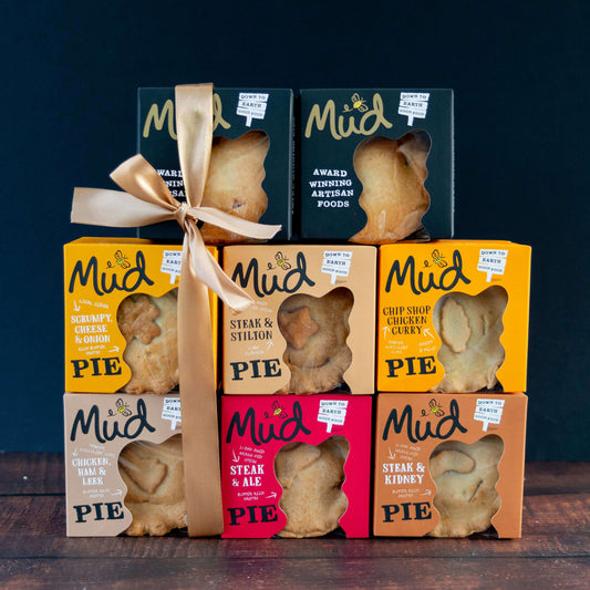 The Ultimate Variety Gift Box from Mud Pies presents seven heart-shaped steak, chicken, and pork pies in vibrant windowed boxes, stacked and finished with a gold ribbon on a dark backdrop.
