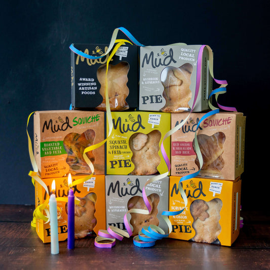 Mud Pies’ Best Ever Veggie Gift Box: savoury pies in vibrant boxes tied with yellow, blue, and pink ribbons, with white and purple candles in front. Squash and mushroom flavours are featured.