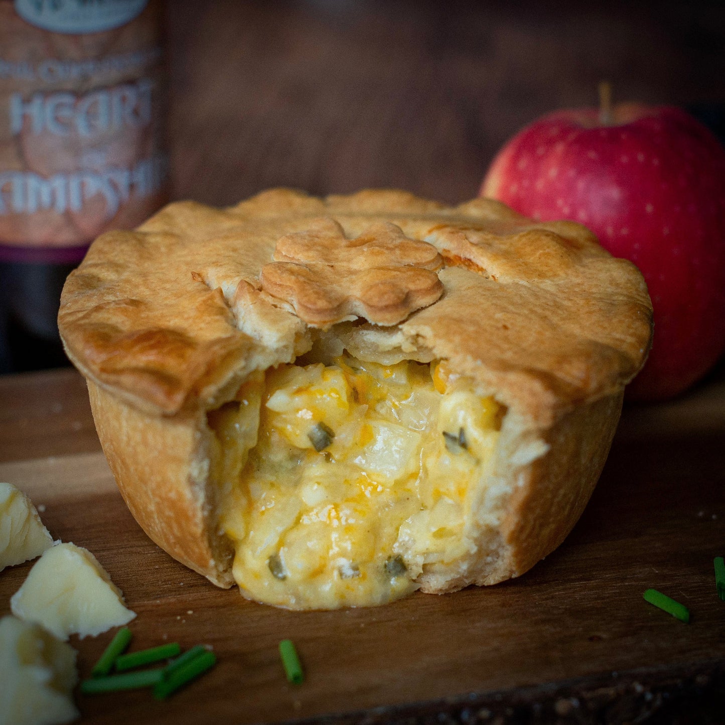 The Mud Pies Best Ever Vegetarian Christmas Gift Box: golden veggie pie, cheese cubes, chives, and red apple on wood.