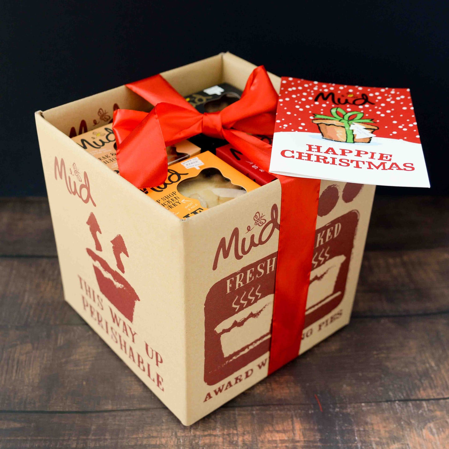 The Ultimate Variety Christmas Gift Box by Mud Pies shows a red-ribboned box, festive bakes, and a “Happie Christmas” card.