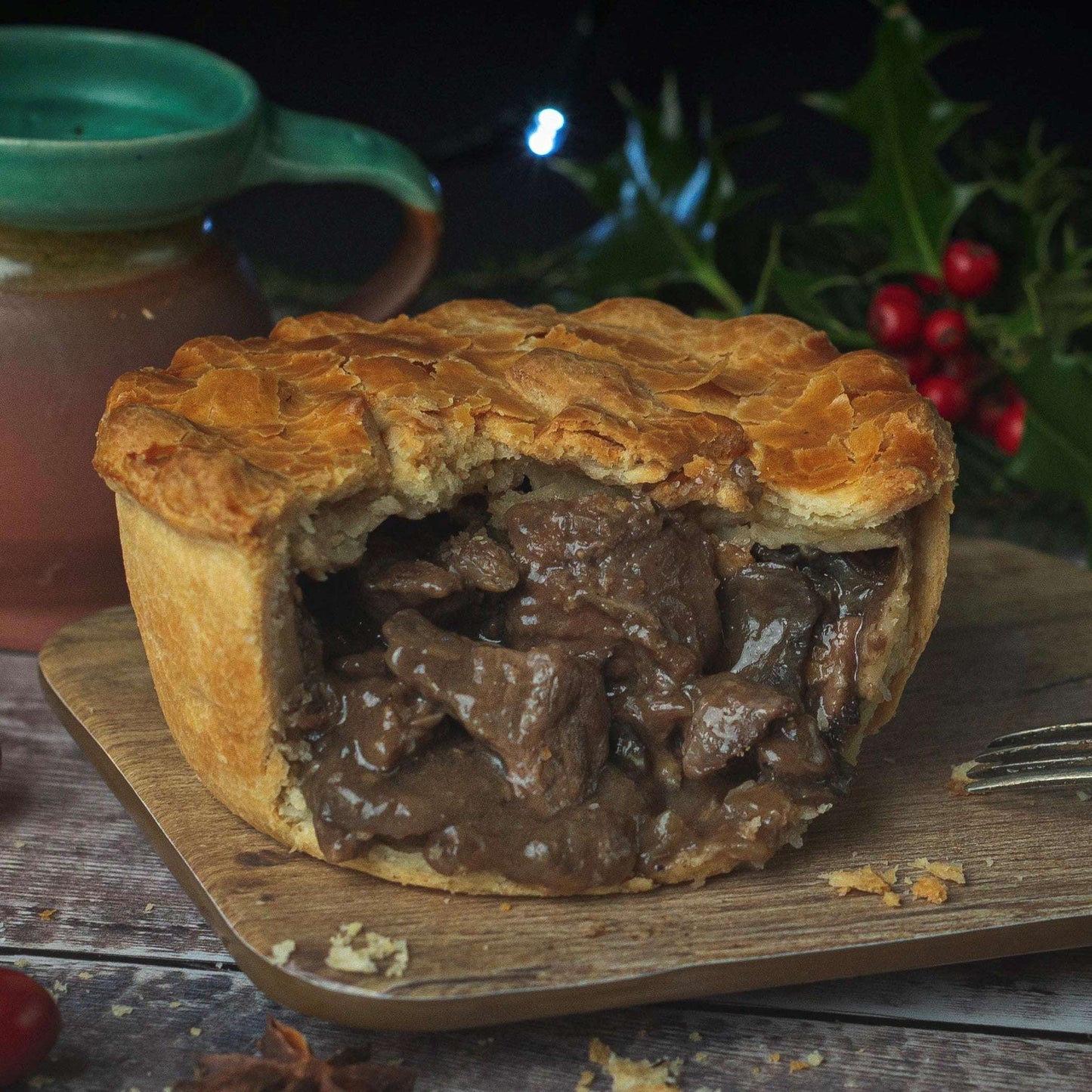 Mud Pies Luxury Christmas Collection steak and ale pie, golden-brown, on wood board, filled with rich beef gravy; mug and holly nearby.