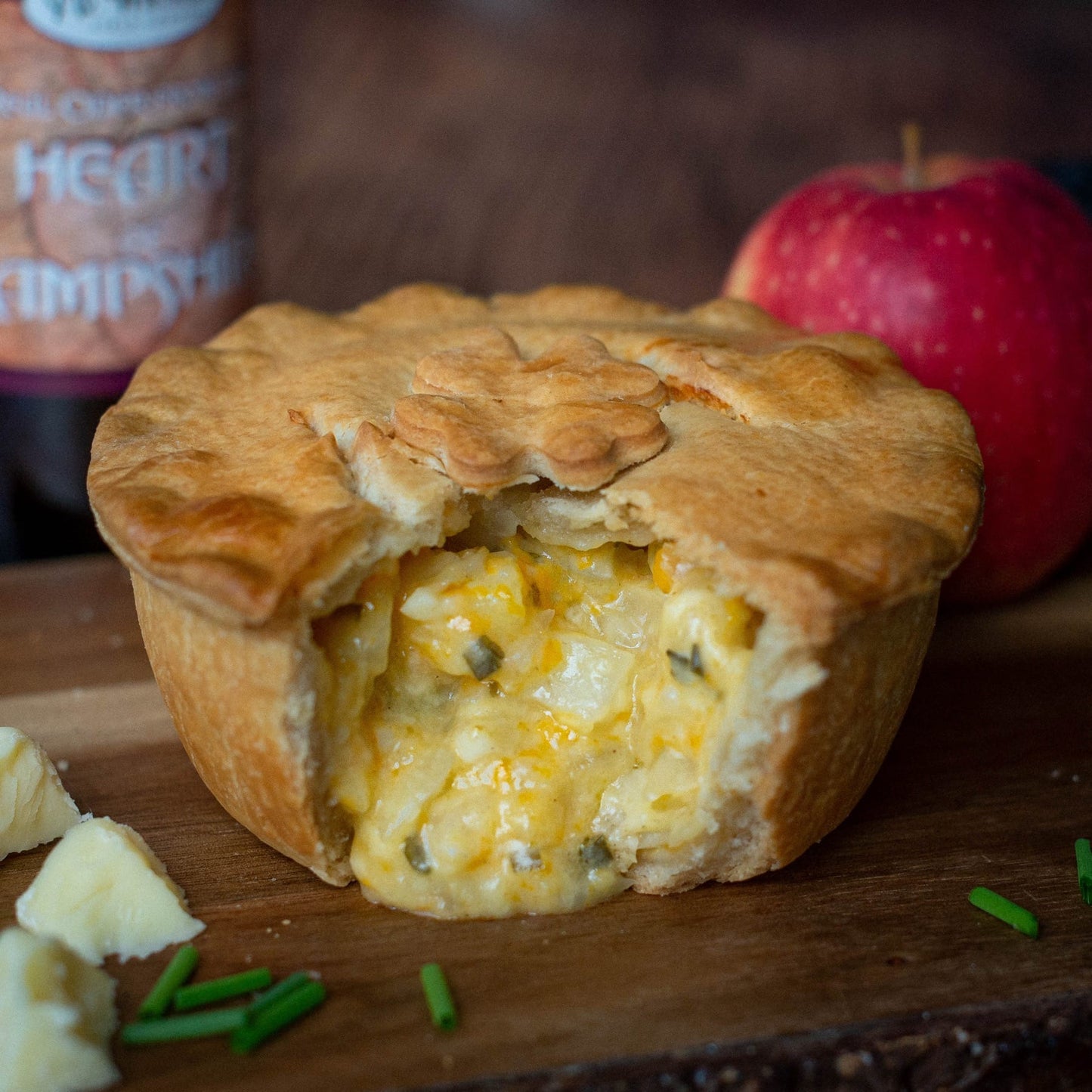 The Mud Pies Scrumpy Cheese and Onion Pie (Cheese, Onion and Potato Pie), golden pastry and creamy filling revealed, sits on a board with chives, cheese, apple.