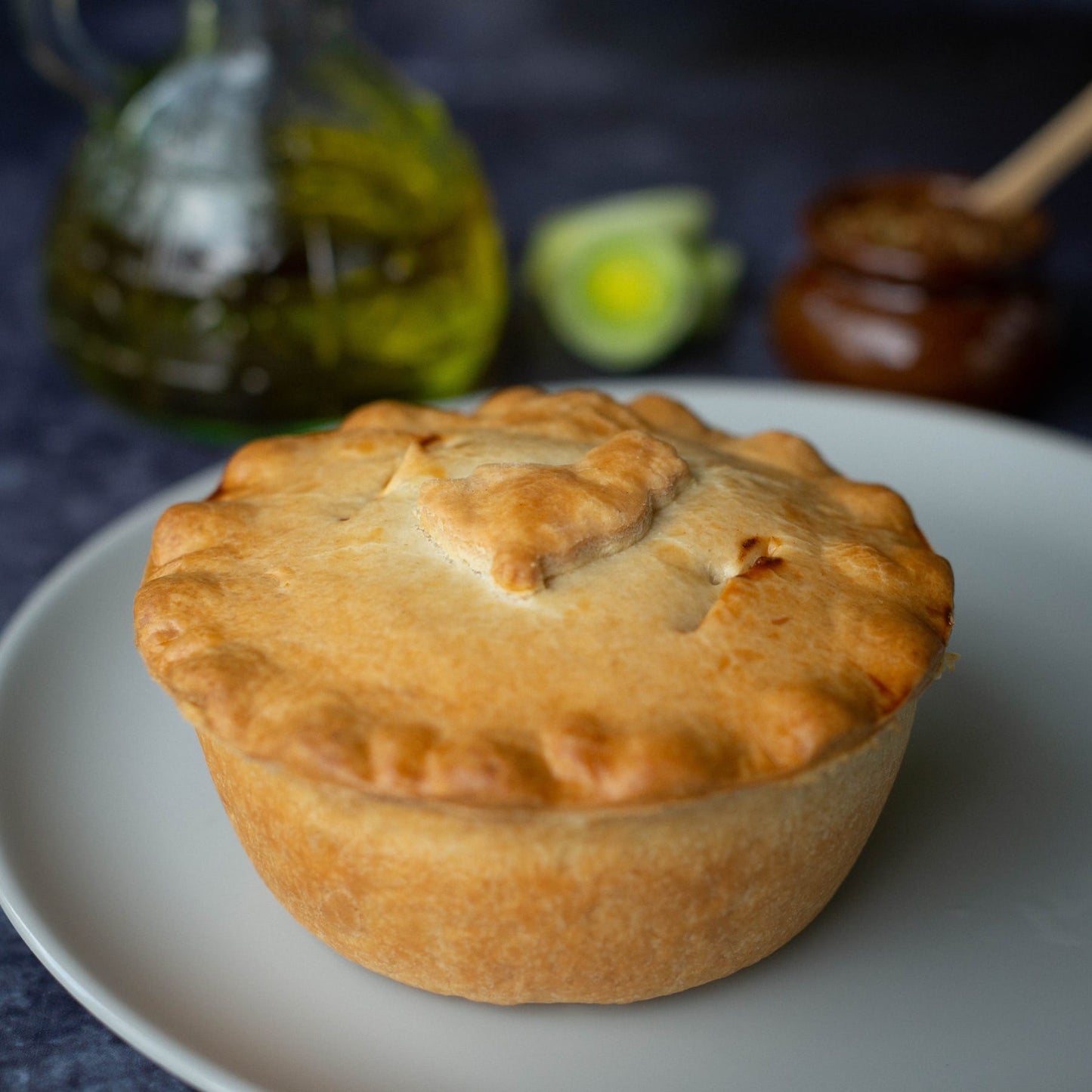 Chicken, Ham and Leek Pie from Mud Pies—golden-brown with buttery shortcrust—rests on a white plate; olive oil and leeks behind.
