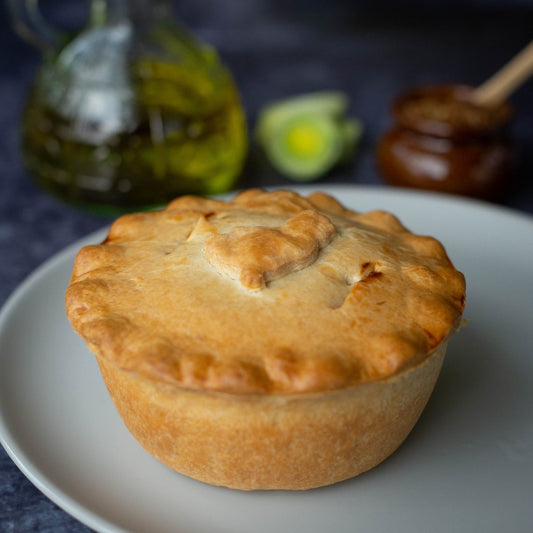 Chicken, Ham and Leek Pie from Mud Pies—golden-brown with buttery shortcrust—rests on a white plate; olive oil and leeks behind.