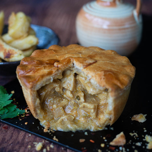 Mud Pies Chip Shop Chicken Curry Pie, golden and flaky, sits sliced open to reveal rich filling; potato wedges and a jug behind.