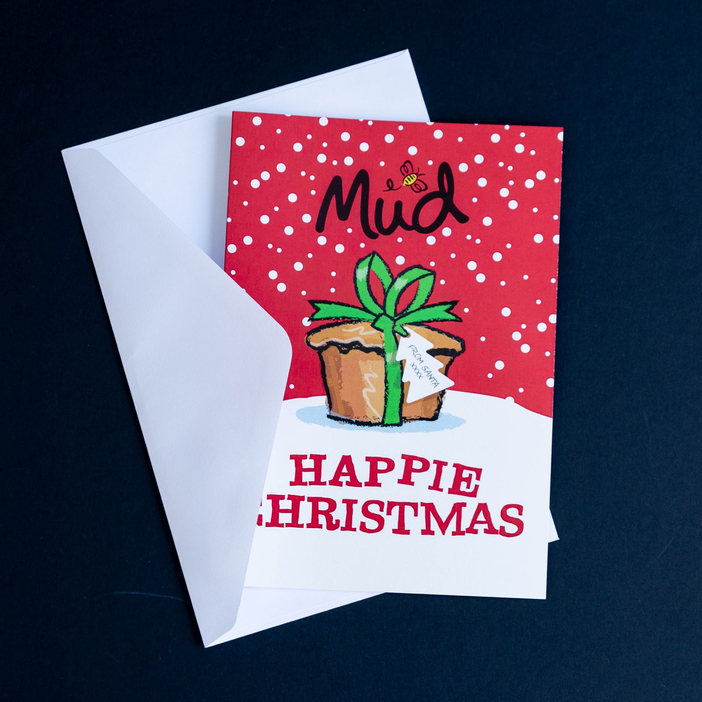 Mud Pies Christmas Greetings Card, tucked in a white envelope on a dark surface, features red snowy trim, pudding motif, bold "HAPPIE CHRISTMAS".