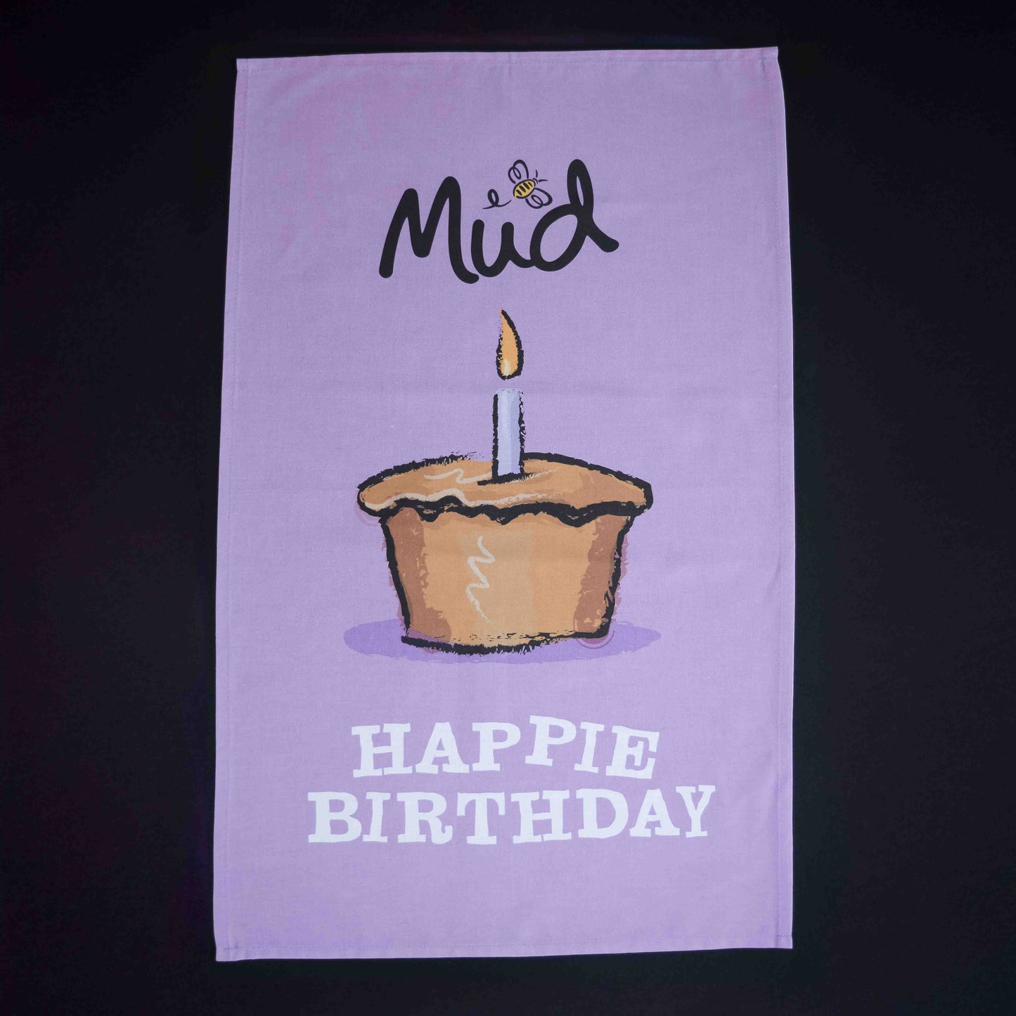 The Mud Pies Tea Towel Birthday is a purple cotton towel with a candle-topped cupcake, bee, “Mud”, and “HAPPIE BIRTHDAY” text.