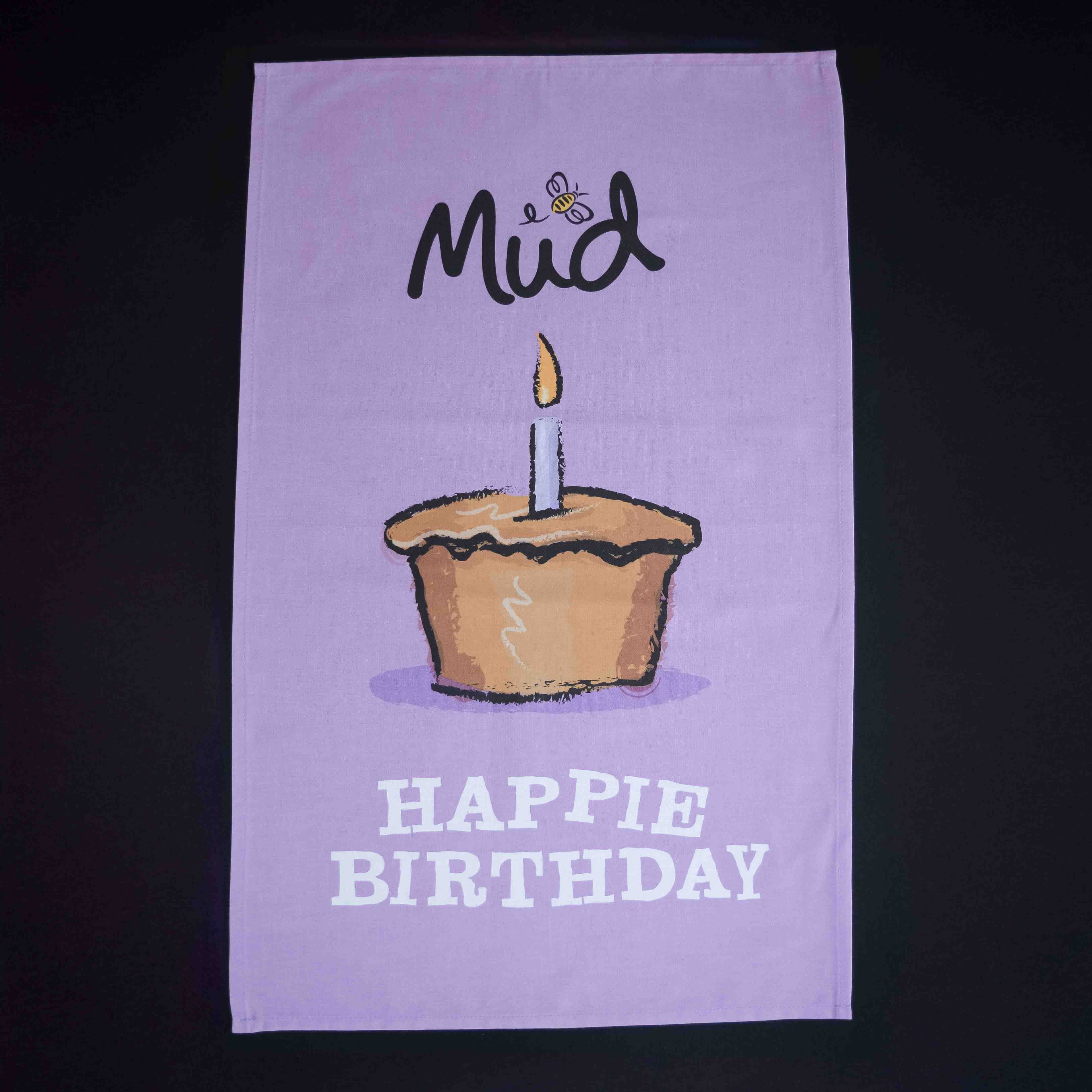 The Mud Pies Tea Towel Birthday is a purple cotton towel with a candle-topped cupcake, bee, “Mud”, and “HAPPIE BIRTHDAY” text.