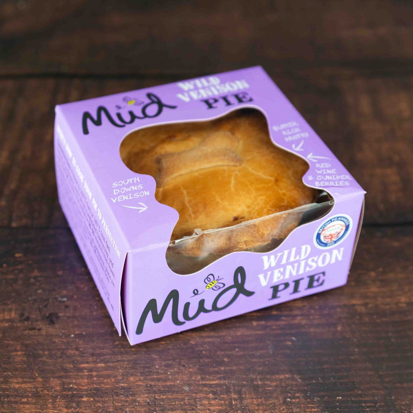 Mud Pies Wild Venison in Red Wine Gravy rests boxed on a dark wood table; golden pie seen through the purple box’s window, with quality award.