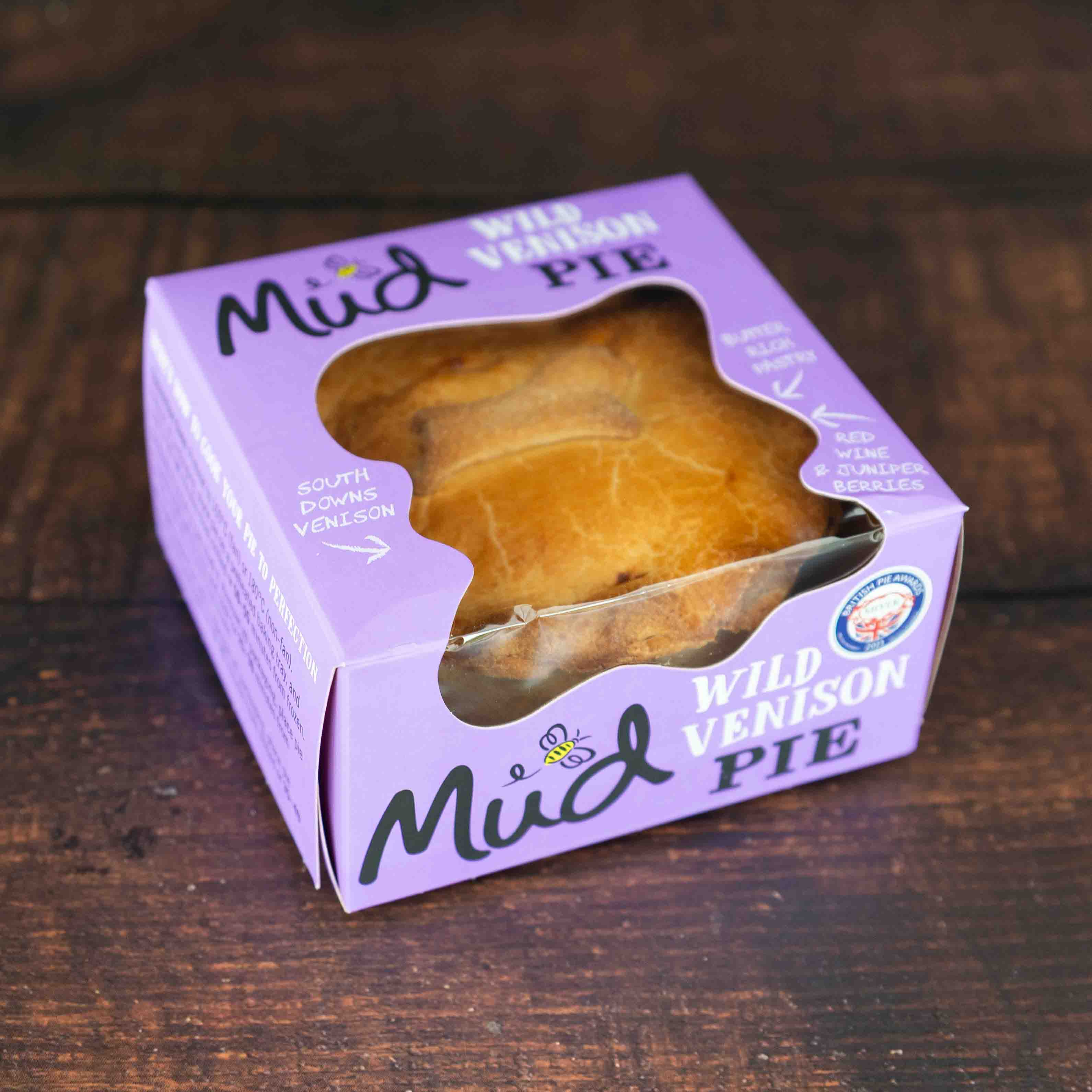Mud Pies Wild Venison in Red Wine Gravy rests boxed on a dark wood table; golden pie seen through the purple box’s window, with quality award.