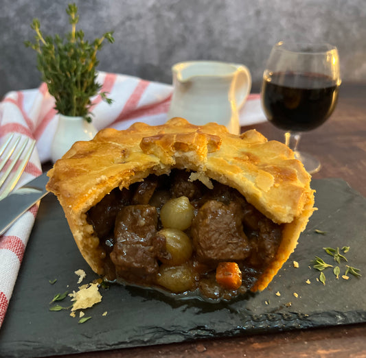 Cut open meat pie on a slate board showing chunks of tender steak, pearl onions and carrots in a beef bourguignon sauce, with a glass of red wine in the background and thyme garnish.