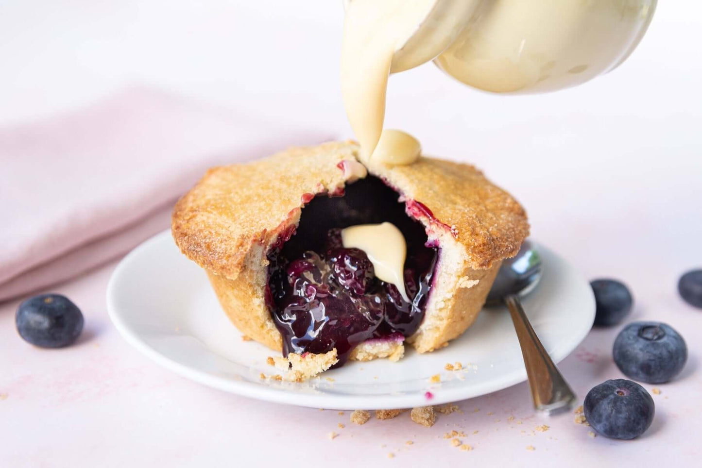 Mud Pies Blueberry Pie, with buttery shortcrust pastry and glossy blueberries, on a white plate as cream is poured over. Blueberries and a spoon rest nearby on the pale pink surface.