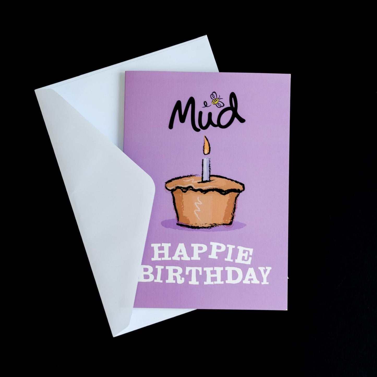 Birthday Greetings Card by Mud Pies, partially beneath a white envelope, shows a lit cupcake, "Mud" with a bee, and bold "HAPPIE BIRTHDAY".