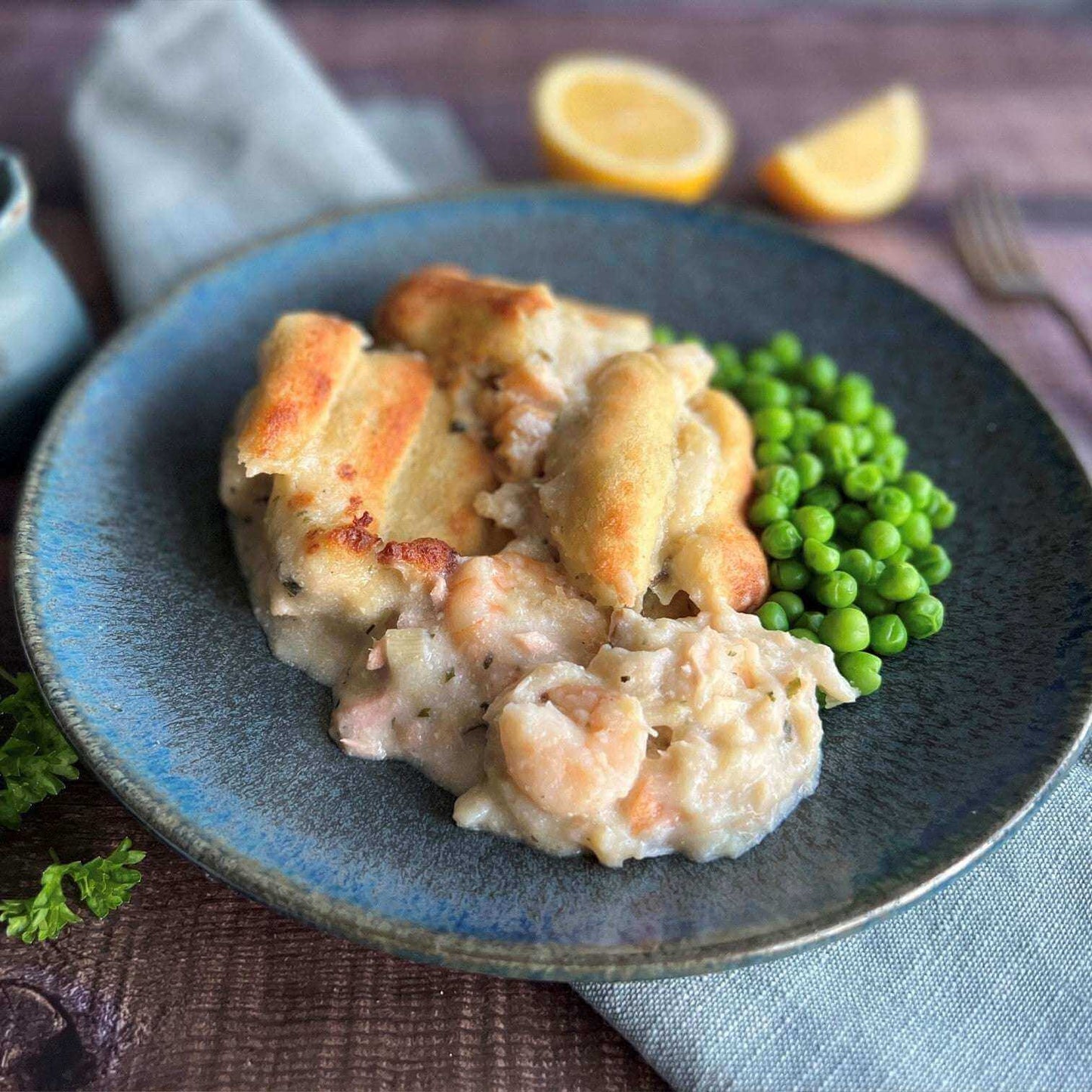 Mud Pies Luxury Fish Pie sits on a blue plate, golden-topped with seafood showing, served beside vibrant peas and lemon halves.