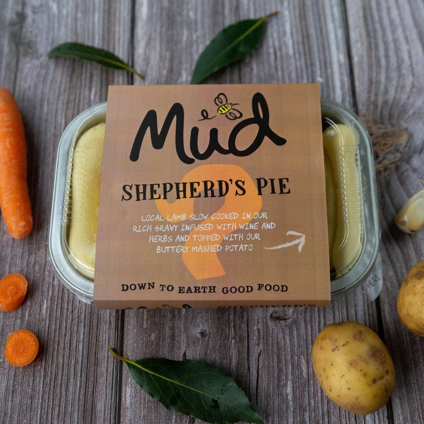 Mud Pies Shepherd's Pie sits on a rustic wood table with fresh carrots, potatoes, bay leaves and a bee-branded logo on the box.