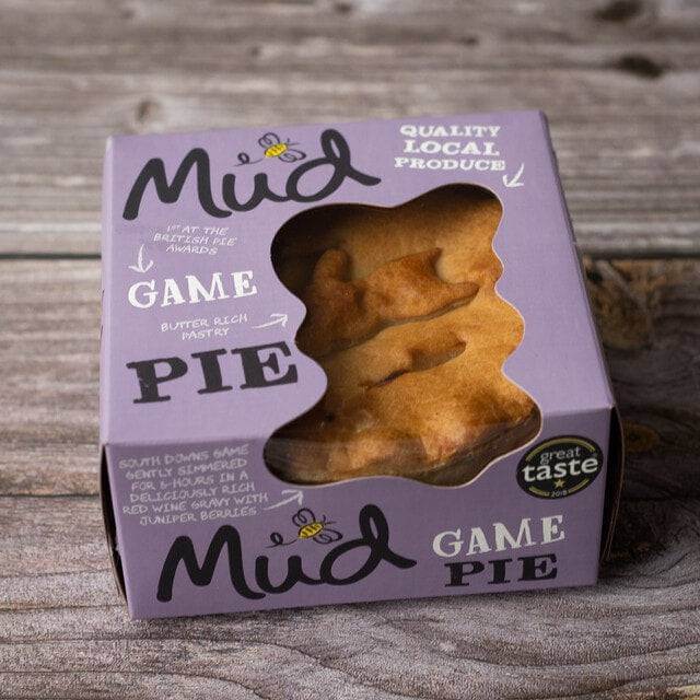 Mud Pies Game Pie sits boxed on wood; the purple box displays a golden pastry topped with birds, local ingredients, and an award.