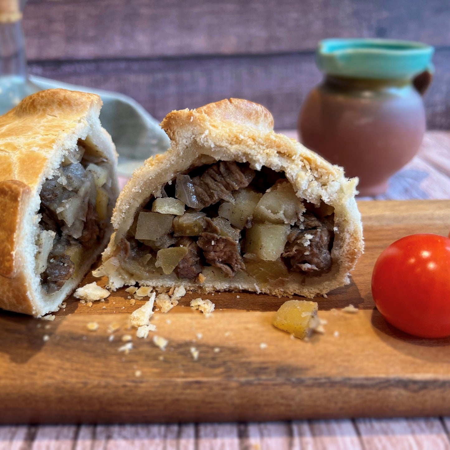 The Mud Pies Dinosaur Pasty is a golden and flaky Cornish Pasty, revealing a rich beef, potato and onion filling on a wooden board with a cherry tomato.