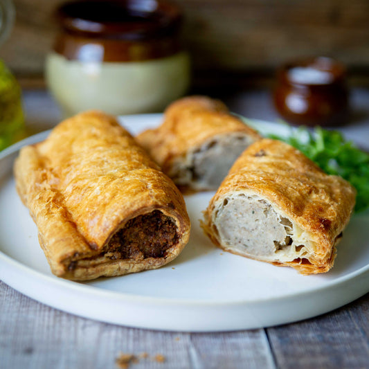 Mud Pies Luxury Sausage Rolls, golden and flaky, rest on a white plate—one whole, one sliced to reveal outdoor-reared pork filling.