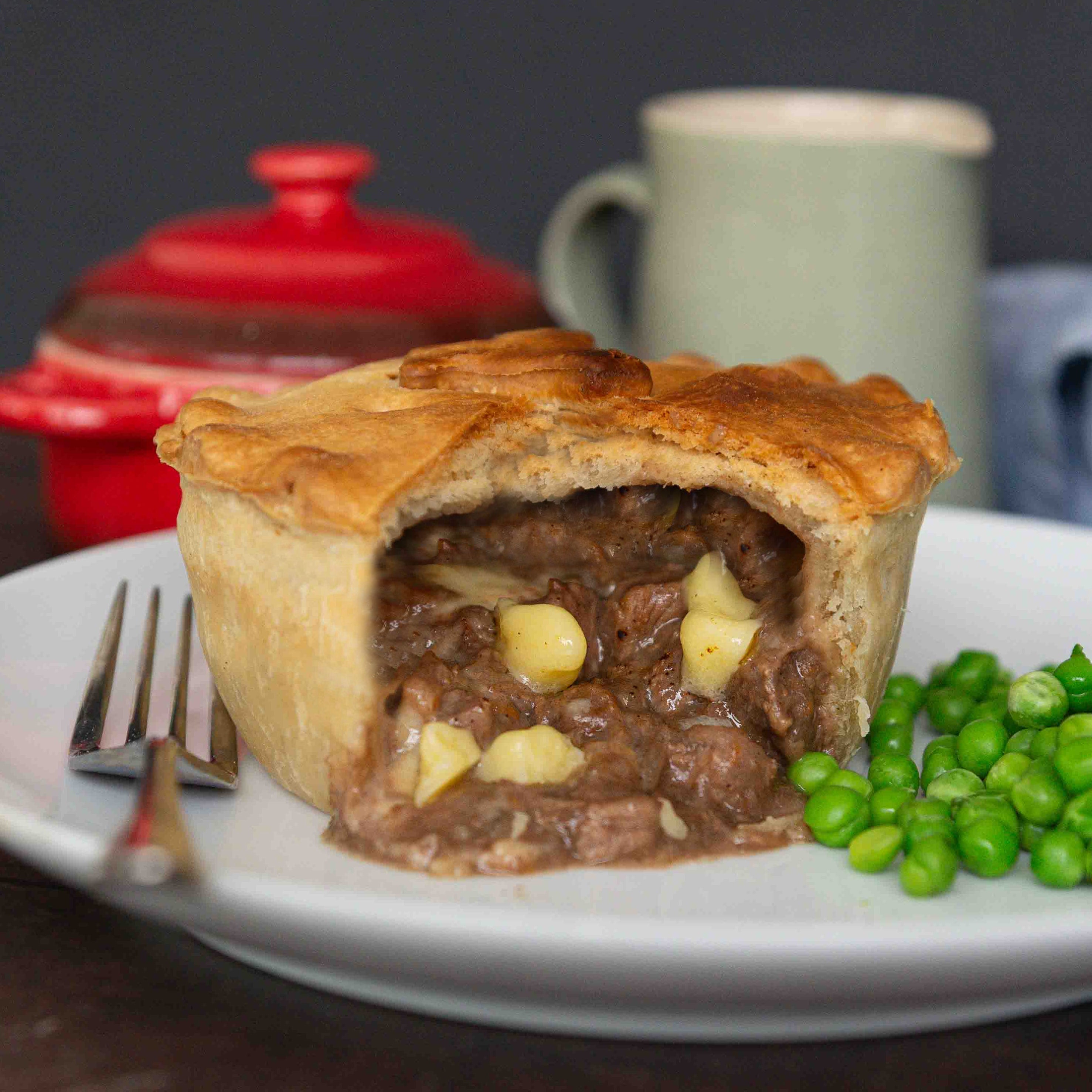 Mud Pies Steak and Cheddar Cheese Pie with a thick, golden crust is sliced open on a white plate, served with green peas.