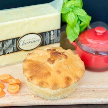 Mud Pies' Steak and Cheddar Cheese Pie, finished with a pastry animal, rests on a wooden board among carrots, cheese, and basil.