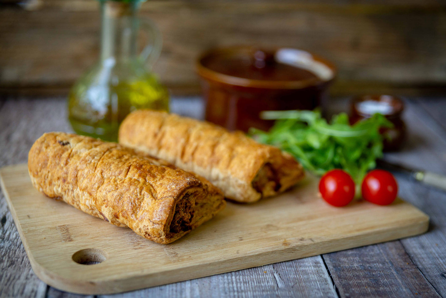 Mud Pies Luxury Sausage Rolls, golden-brown and flaky, rest on a wooden board with fresh greens and cherry tomatoes; rustic items behind.