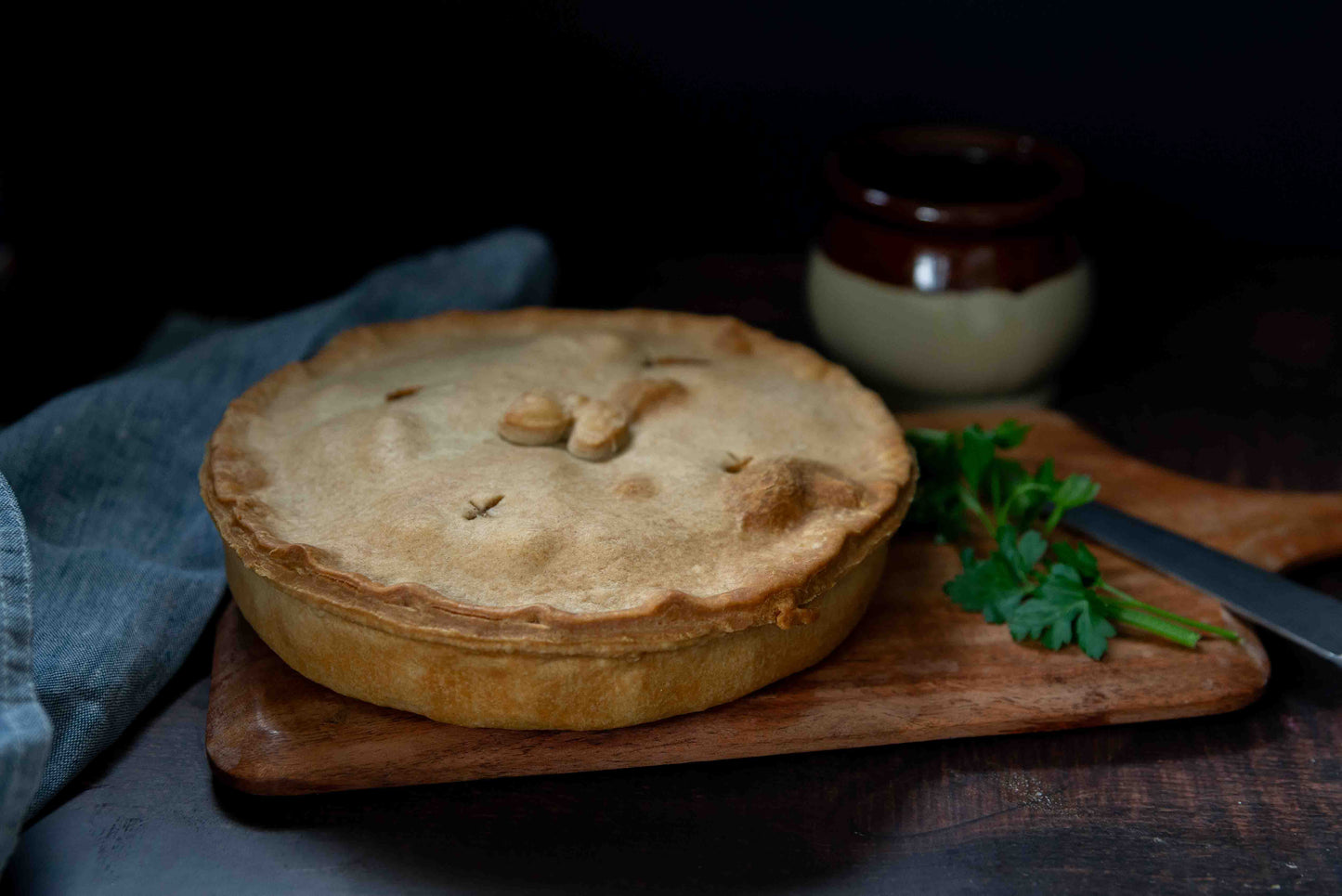 Mud Pies' Steak and Kidney Family Pie on a wooden board with parsley, knife, ceramic pot and blue cloth in the background.