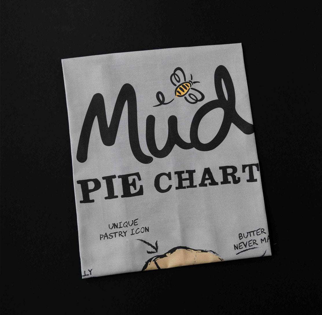 Tea Towel Pie Chart by Mud Pies—folded grey 100 cotton, on black surface, with “Mud PIE CHART”, bee and pie graphics.
