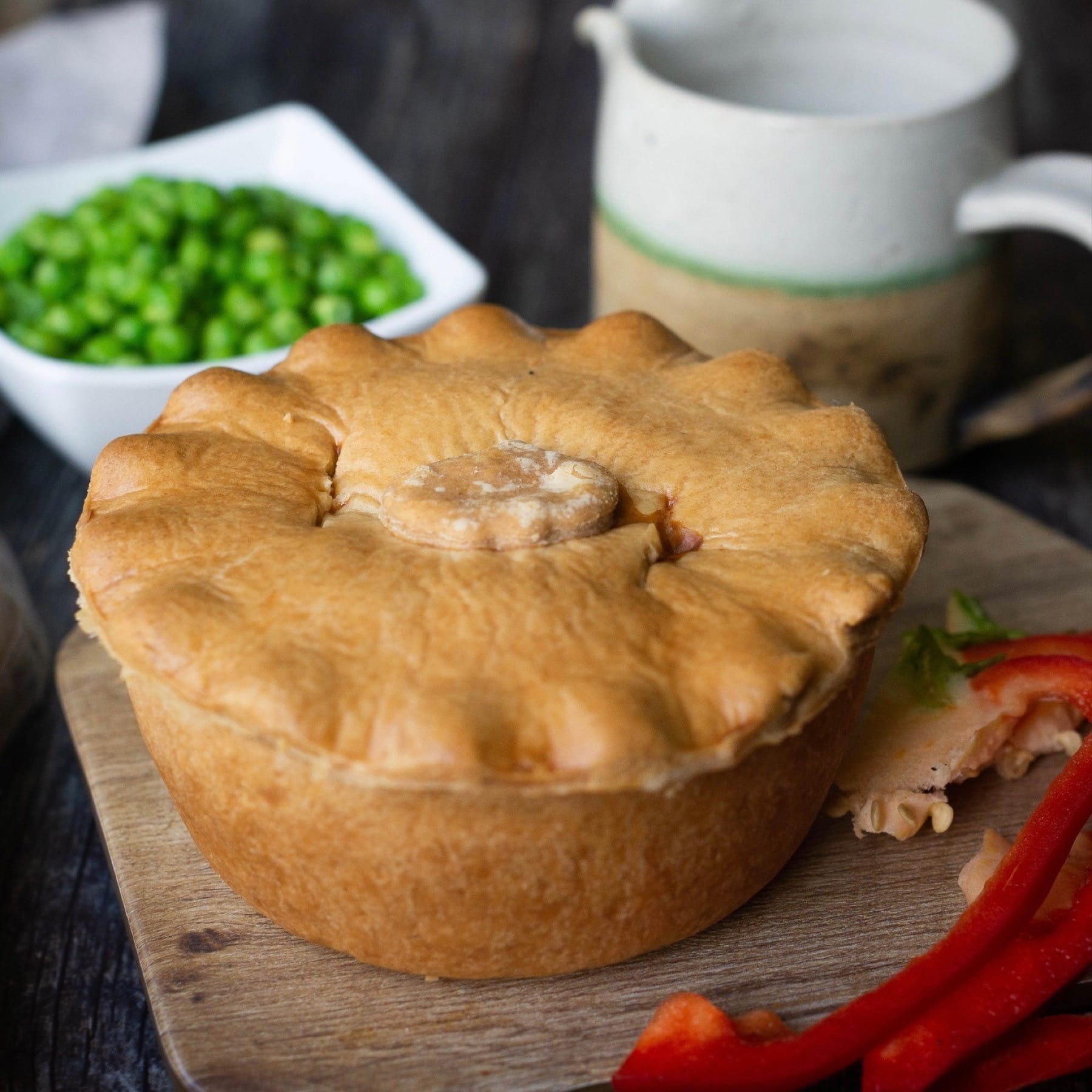 The Mud Pies Squash, Spinach and Feta Pie vegan is golden-brown with crimped edges, on a board with peppers and veg.
