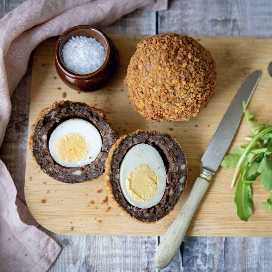 Mud Pies Scotch Egg on a wooden board—one whole, one sliced to reveal free-range egg in sausage crumb. Arugula, salt, knife, and pink napkin nearby.