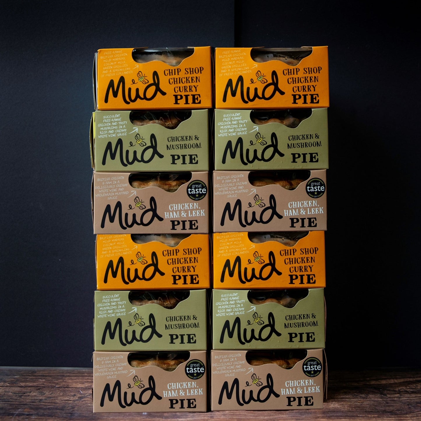 Twelve Mud Pies Chicken Pie Box 12 Pack stacked in two neat columns on wood, showing bestselling pies in orange, brown, and green packs.