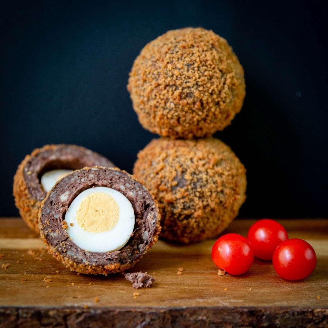 Mud Pies Scotch Egg on a wooden board, halved to show a free-range egg in golden sausage crumb, three cherry tomatoes beside it.