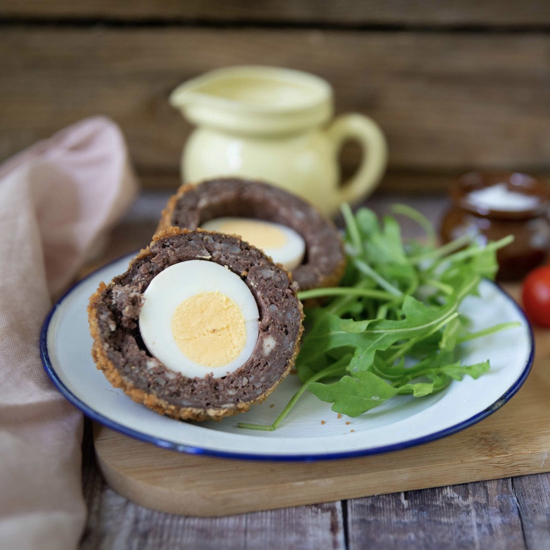 Mud Pies Scotch Egg single: a sliced free-range egg wrapped in sausage and breadcrumbs, on a plate with arugula; pale jars and a yellow jug rest on wooden boards by a pink cloth in the background.