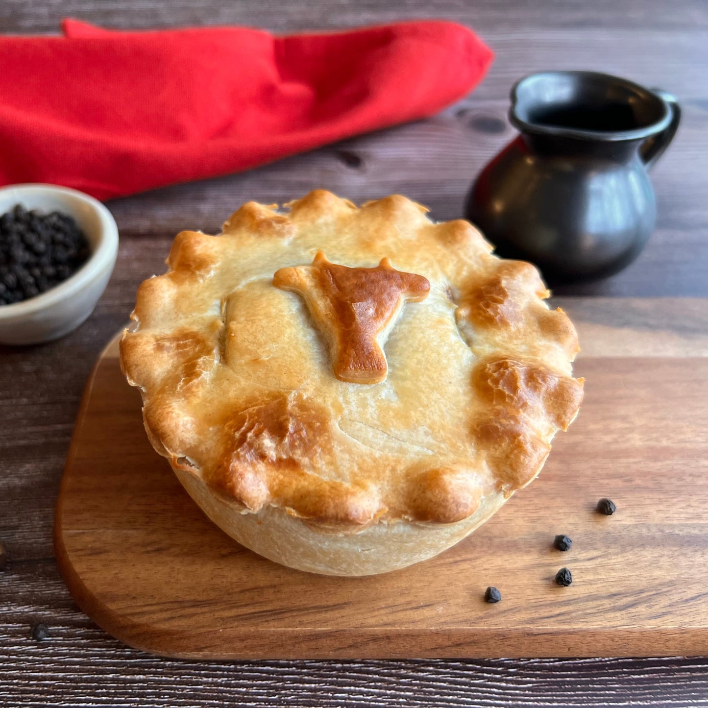 Mud Pies’ Peppered Steak Pie boasts a golden crust with a pastry cow, on a wooden board with peppercorns, pepper bowl, and red napkin.