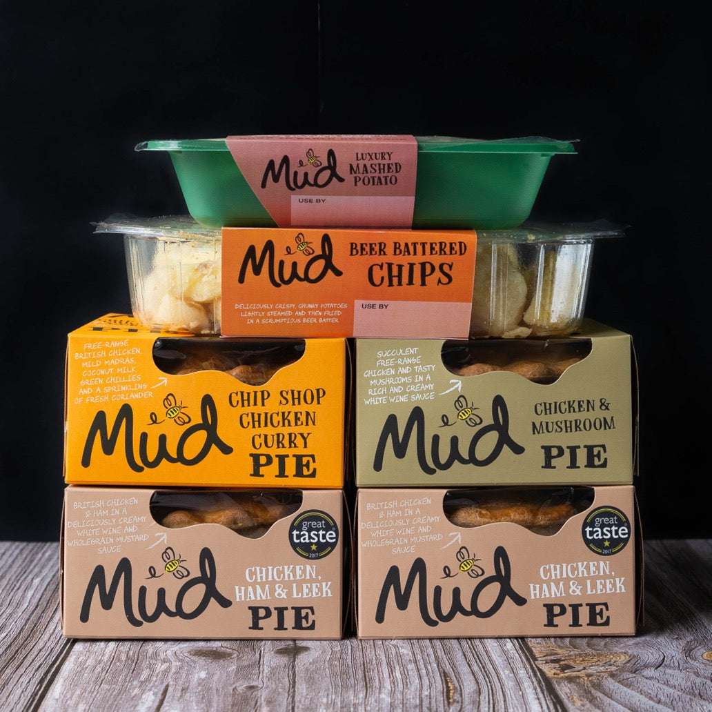 Mud Pies Chicken Dinner tops a neatly stacked selection of their products on a wooden table, set before a deep, dark backdrop.