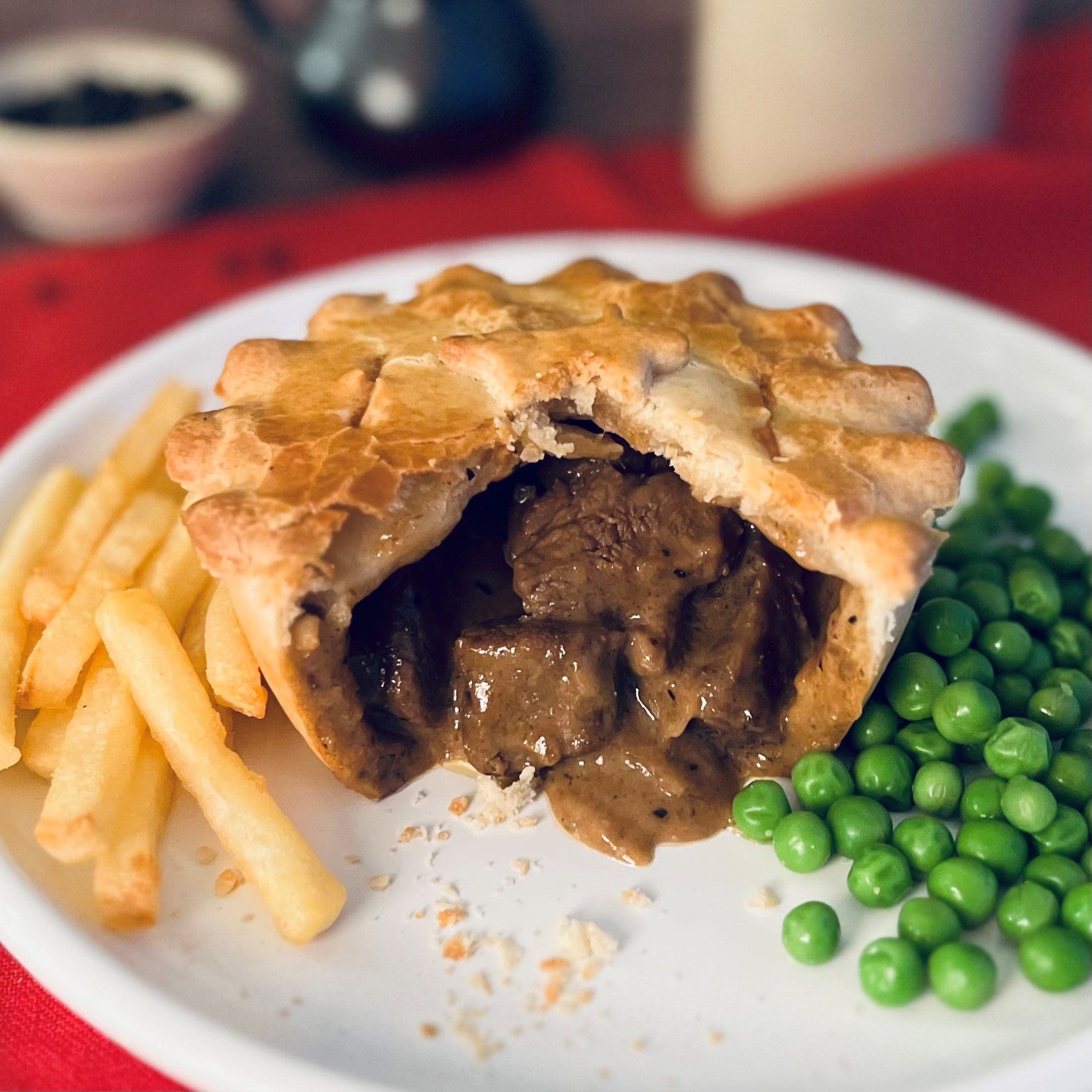 Mud Pies Peppered Steak Pie, golden and half-eaten, reveals rich filling on a white plate with chips and peas over a red tablecloth.
