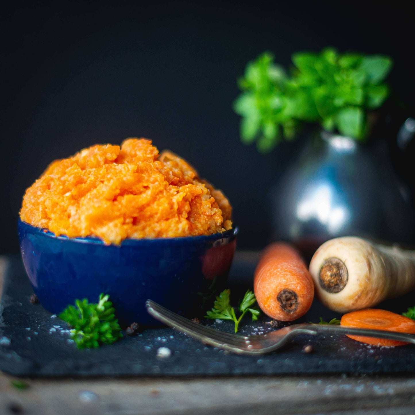 Mud Pies Carrot and Parsnip Mash in a blue bowl on a slate board, with carrot, parsnip, parsley, fork and fresh herbs in view.