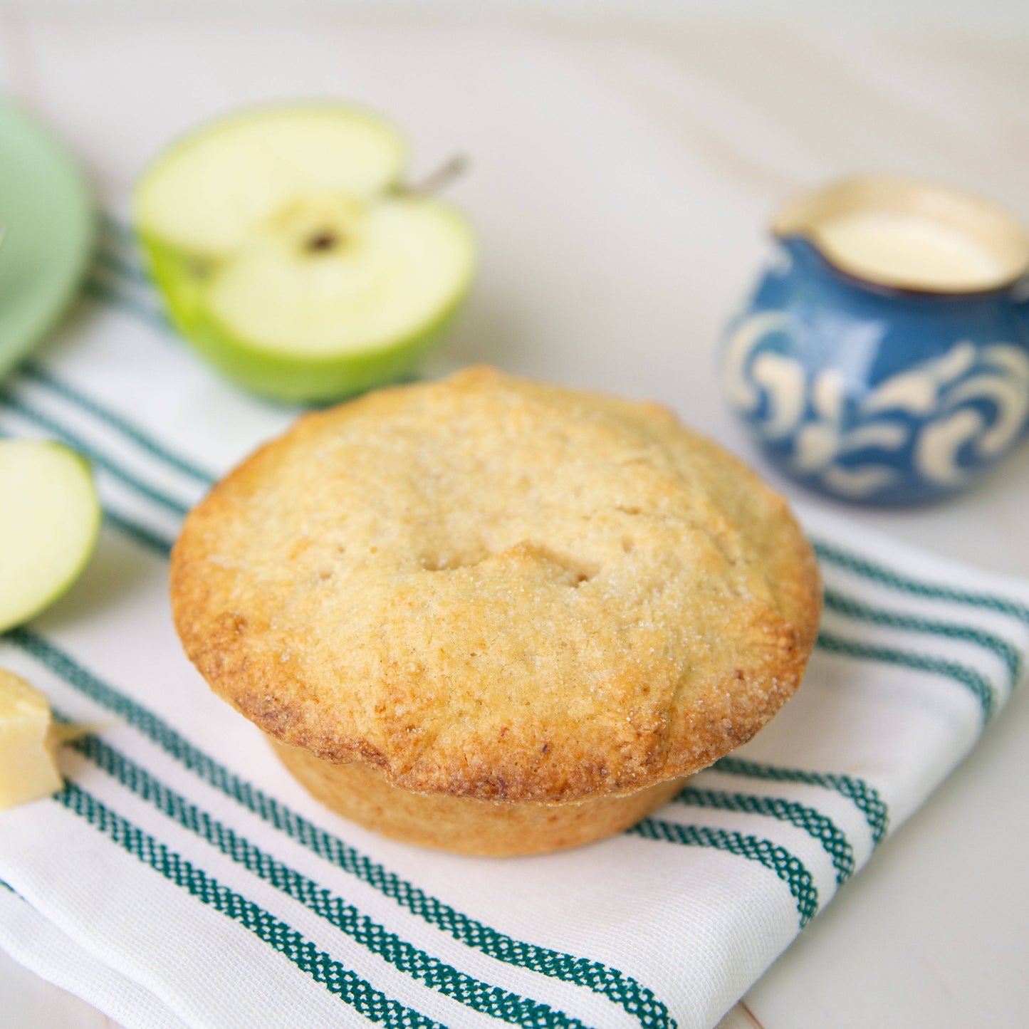 Mud Pies Apple Pie with Cheddar Cheese Crust, golden-brown, sits on a green-striped cloth with apples and a blue-white jug behind.