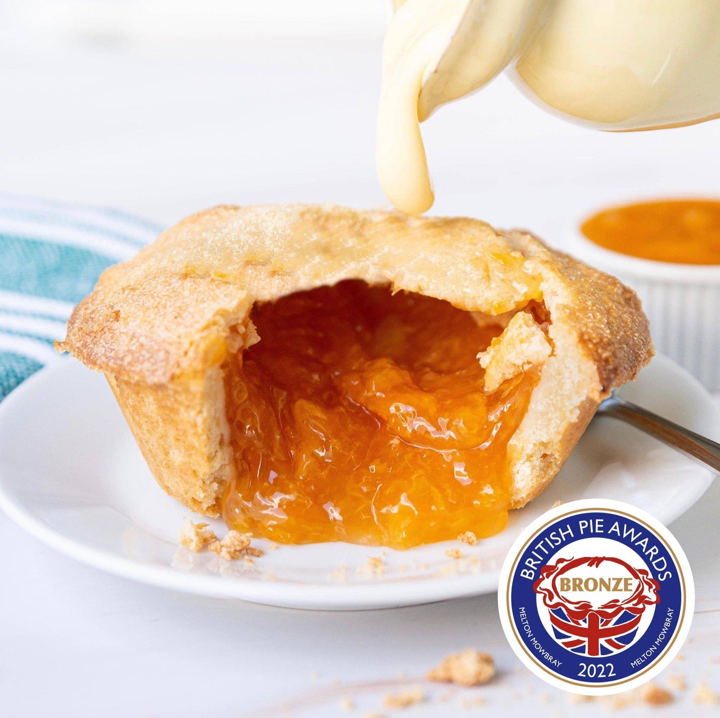 Mud Pies Apricot Pie boasts a golden pastry, vivid apricot filling and crumbs on a white plate, topped with cream and a striped napkin.