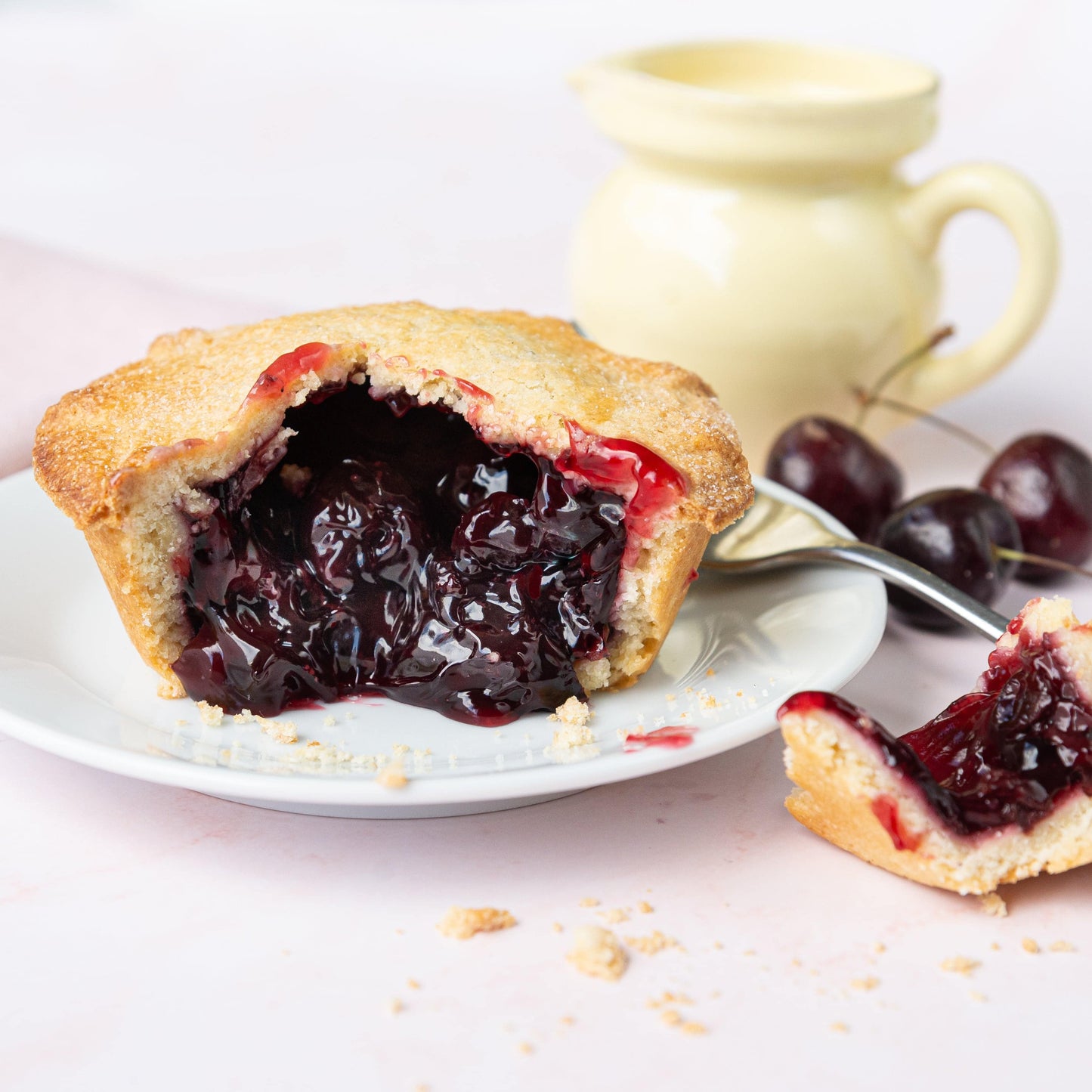 Mud Pies Black Cherry Pie with golden buttery shortcrust pastry on a white plate, cut to show glossy cherries; crumbs nearby, yellow jug with cream and fresh cherries behind on pale pink.