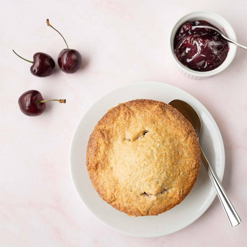 Mud Pies Black Cherry Pie with buttery shortcrust pastry on a white plate, spoon beside, two dark cherries and cherry jam nearby.