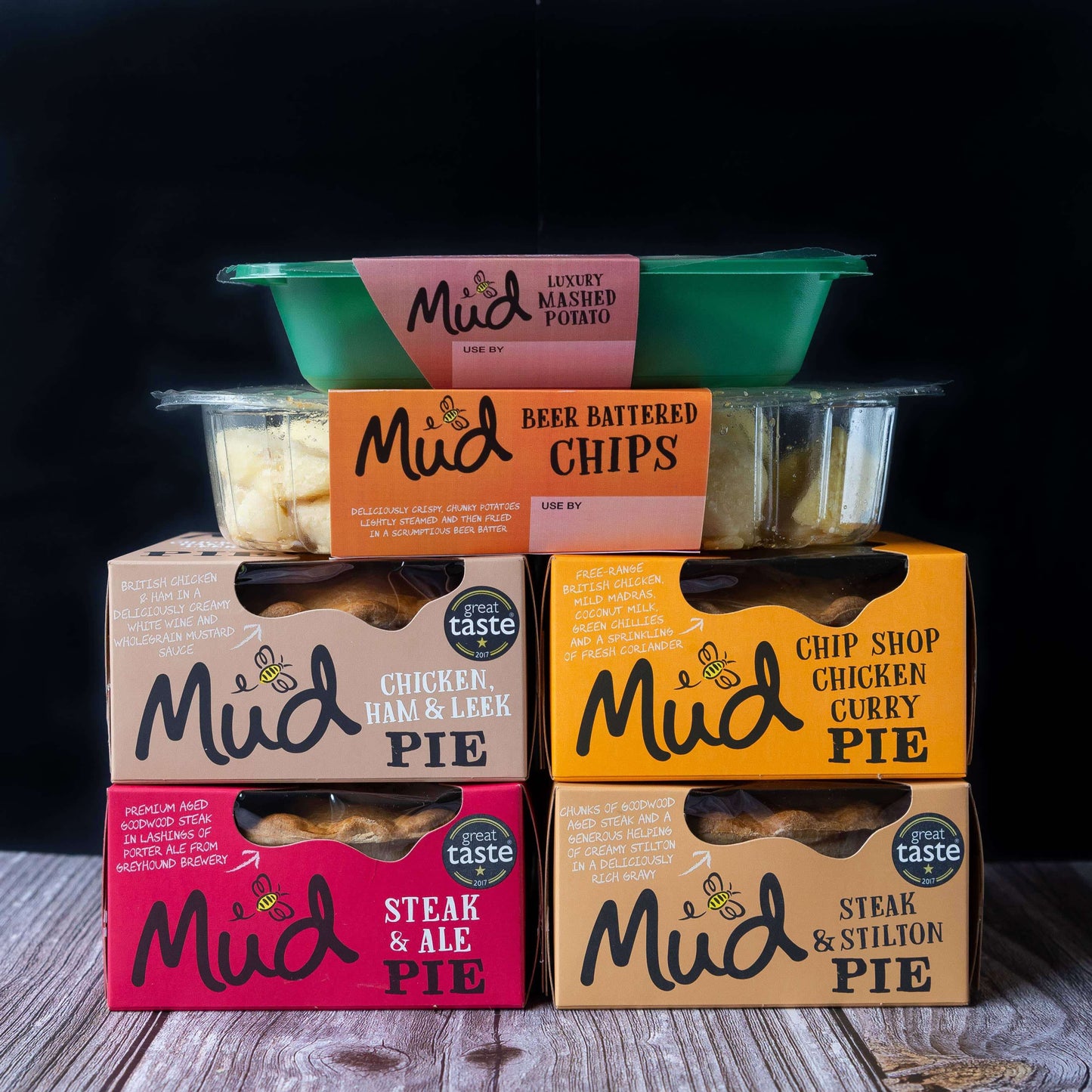 The Mud Pies Best Sellers Dinner displays five boxed pies in classic flavours, with beer-battered chips and creamy mash, neatly stacked on wood.