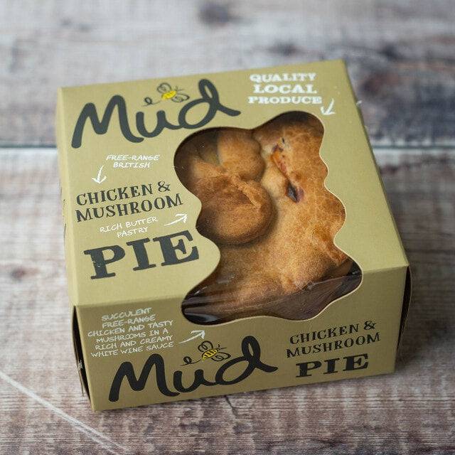 Mud Pies Chicken Pie, in earthy green packaging, rests on a wooden table; a window shows its golden crust and quality local ingredients.