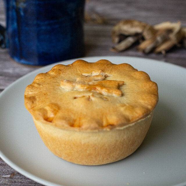 Mud Pies Game Pie, golden-brown with a crimped edge and pastry leaf, sits on a white plate; blue mug and mushrooms behind.