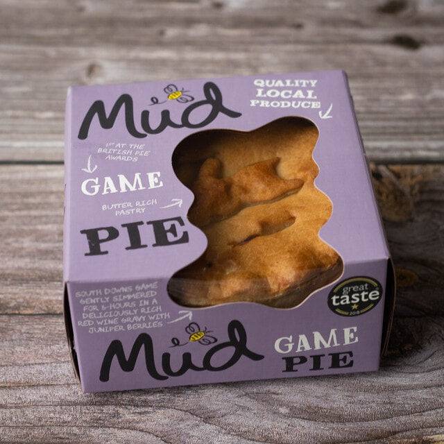Mud Pies Game Pie sits boxed on wood; the purple box displays a golden pastry topped with birds, local ingredients, and an award.
