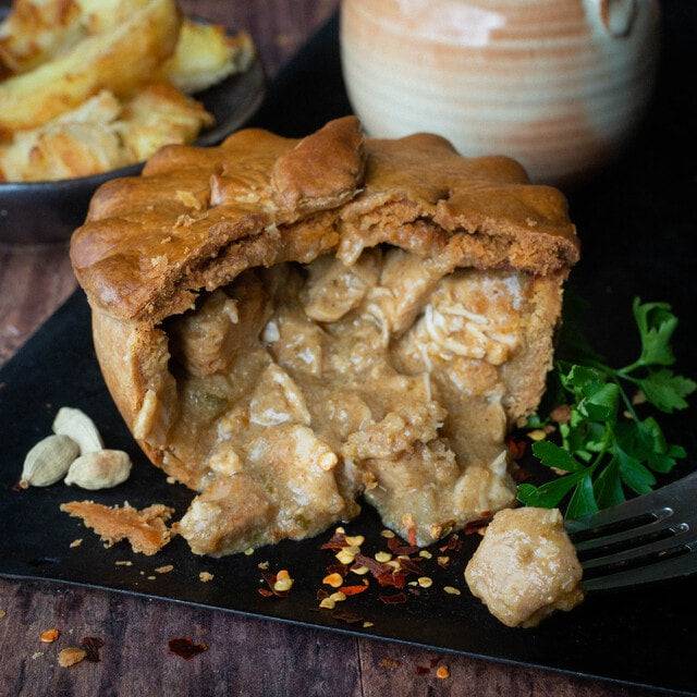The Mud Pies Gluten-Free Chip Shop Chicken Curry Pie, golden and flaky, reveals succulent British chicken in creamy curry; chilli flakes, cardamom pods, parsley and a fork beside it, with potato wedges and a cup in the background.