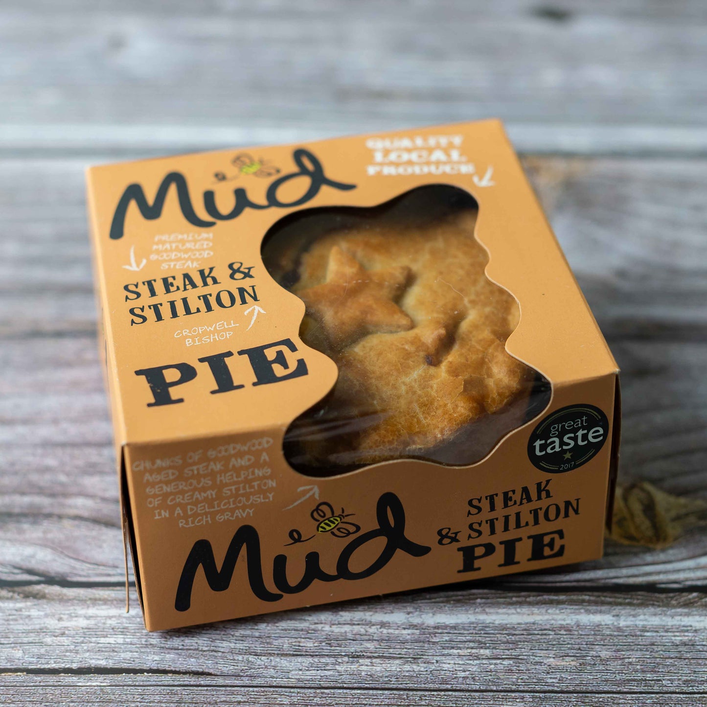 Mud Pies Steak and Stilton Pie sits boxed on aged wood; brown pack with window reveals golden pastry, “great taste” badge top right.