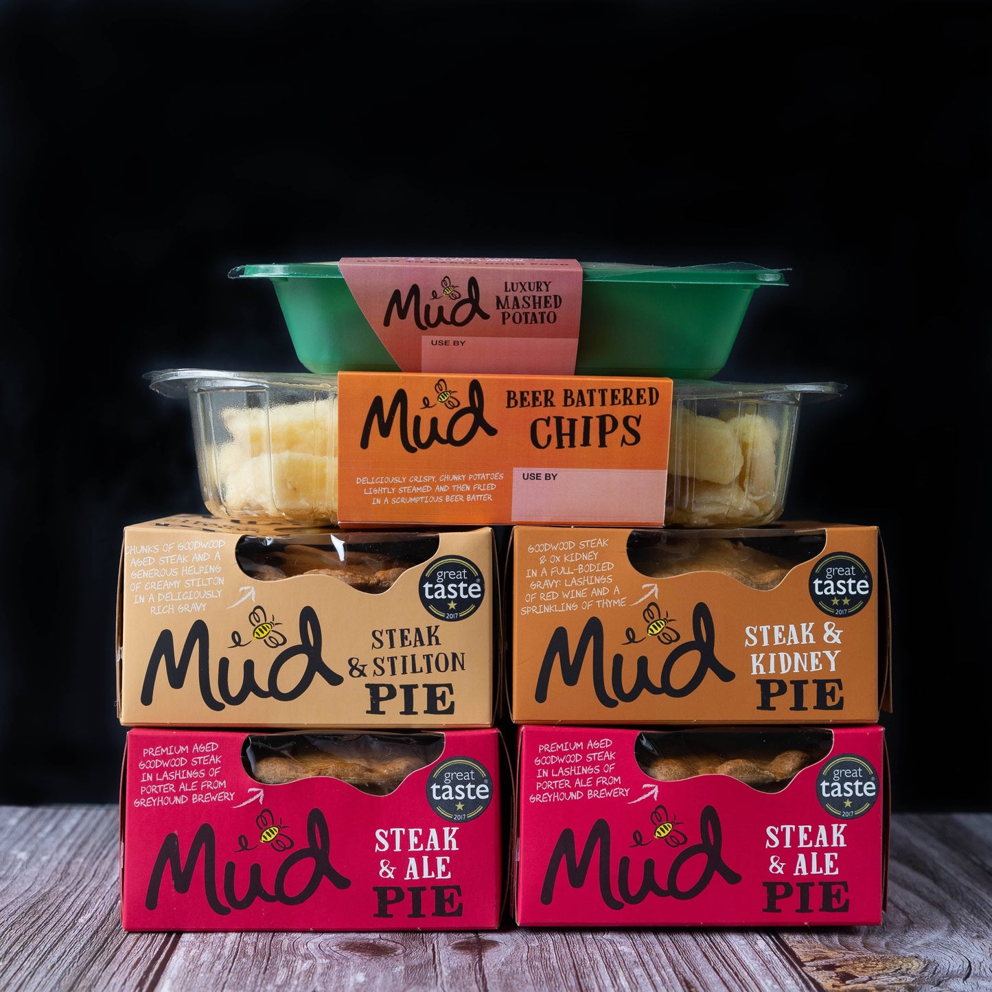 Mud Pies' Steak Dinner meals are stacked neatly on a wooden surface, evoking cosy British comfort against a dark backdrop.
