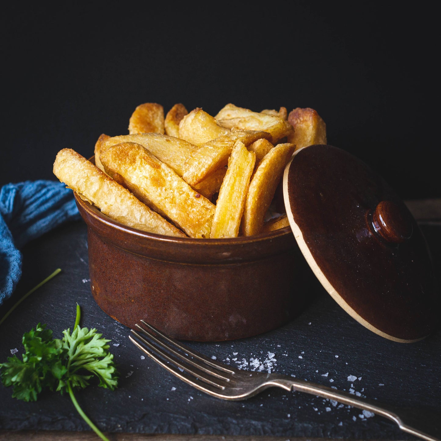 Mud Pies ceramic dish filled with golden Beer Battered Chips sits on dark slate; lid, parsley, sea salt, fork, and blue napkin nearby.