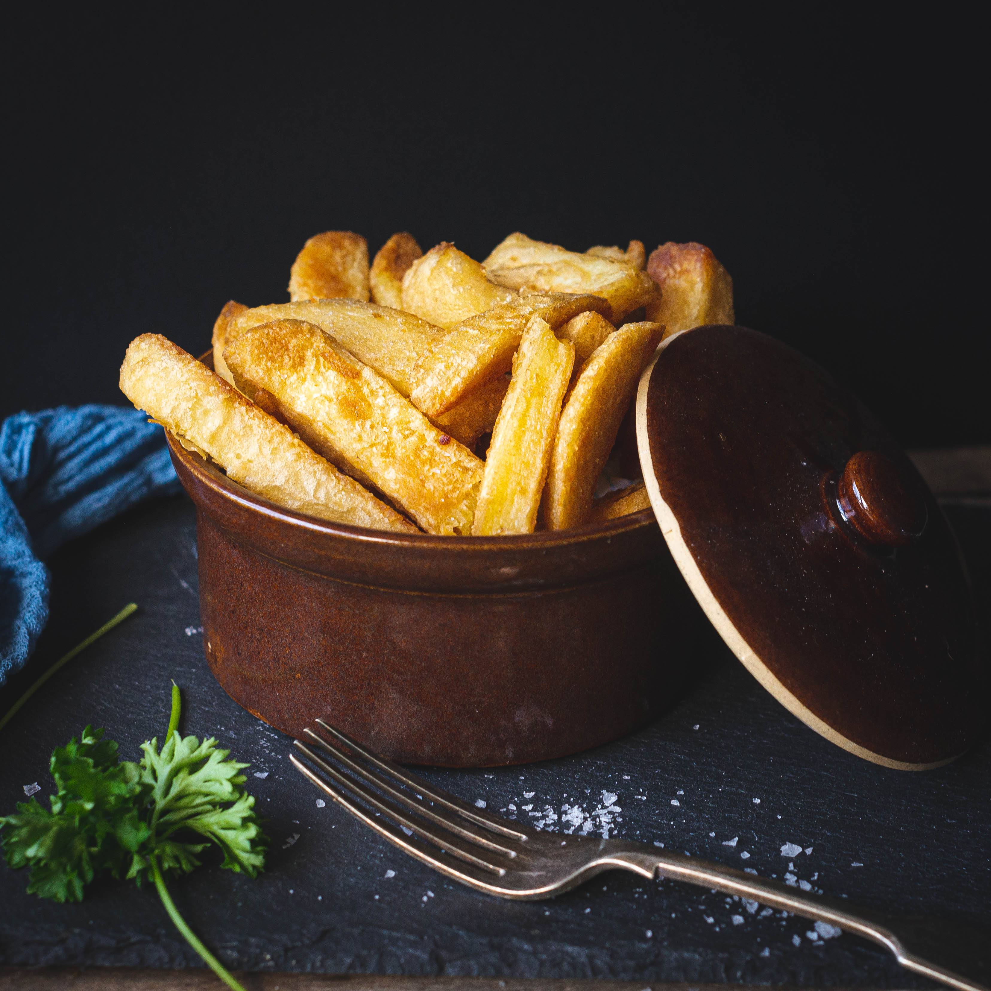 Mud Pies ceramic dish filled with golden Beer Battered Chips sits on dark slate; lid, parsley, sea salt, fork, and blue napkin nearby.