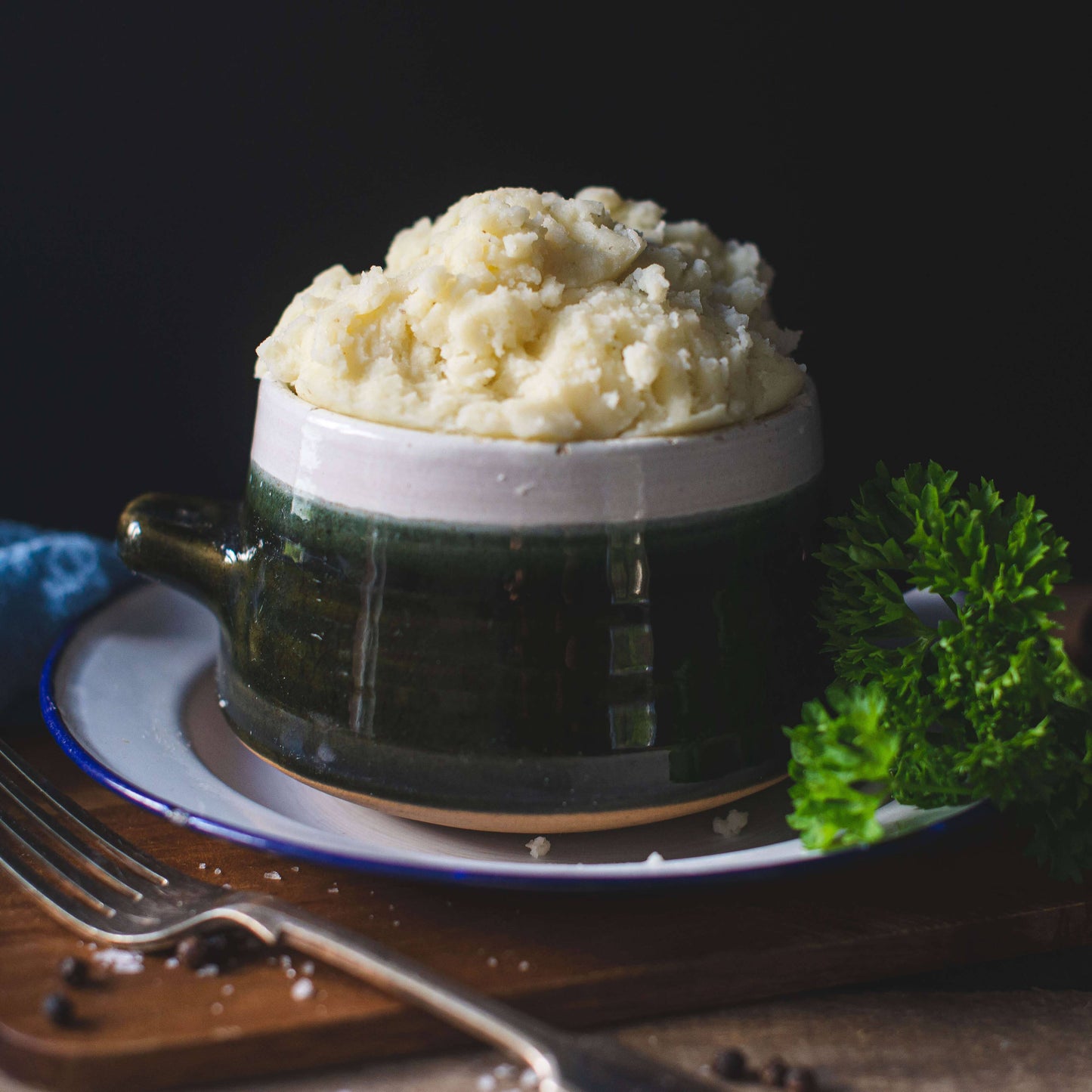 Mud Pies’ Buttery Mashed Potato in a green and white bowl, topped with parsley and peppercorns, sits on a white plate with a fork.